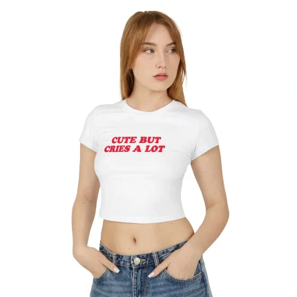 A woman with long light brown hair models the Cute But Cries a Lot Baby Tee, a white crop top featuring red text, paired with blue jeans. She stands against a plain white background, looking to the side with her hands in her pockets.