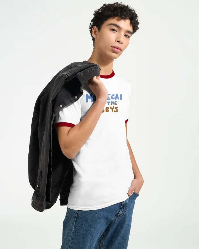 A person with short curly hair wears a Mordecai and the Rigbys Shirt and blue jeans, holding a black jacket over their shoulder, standing against a plain light background.